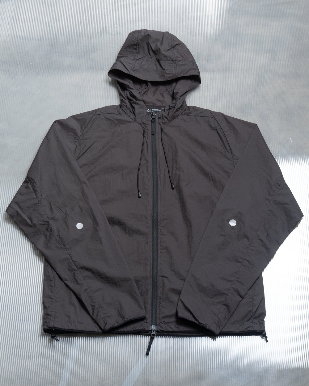 A black nylon taffeta jacket with an adjustable drawstring hood, two-way zip, dot accents, two seam pockets, and thumbholes at the cuff, displayed on a grey background.