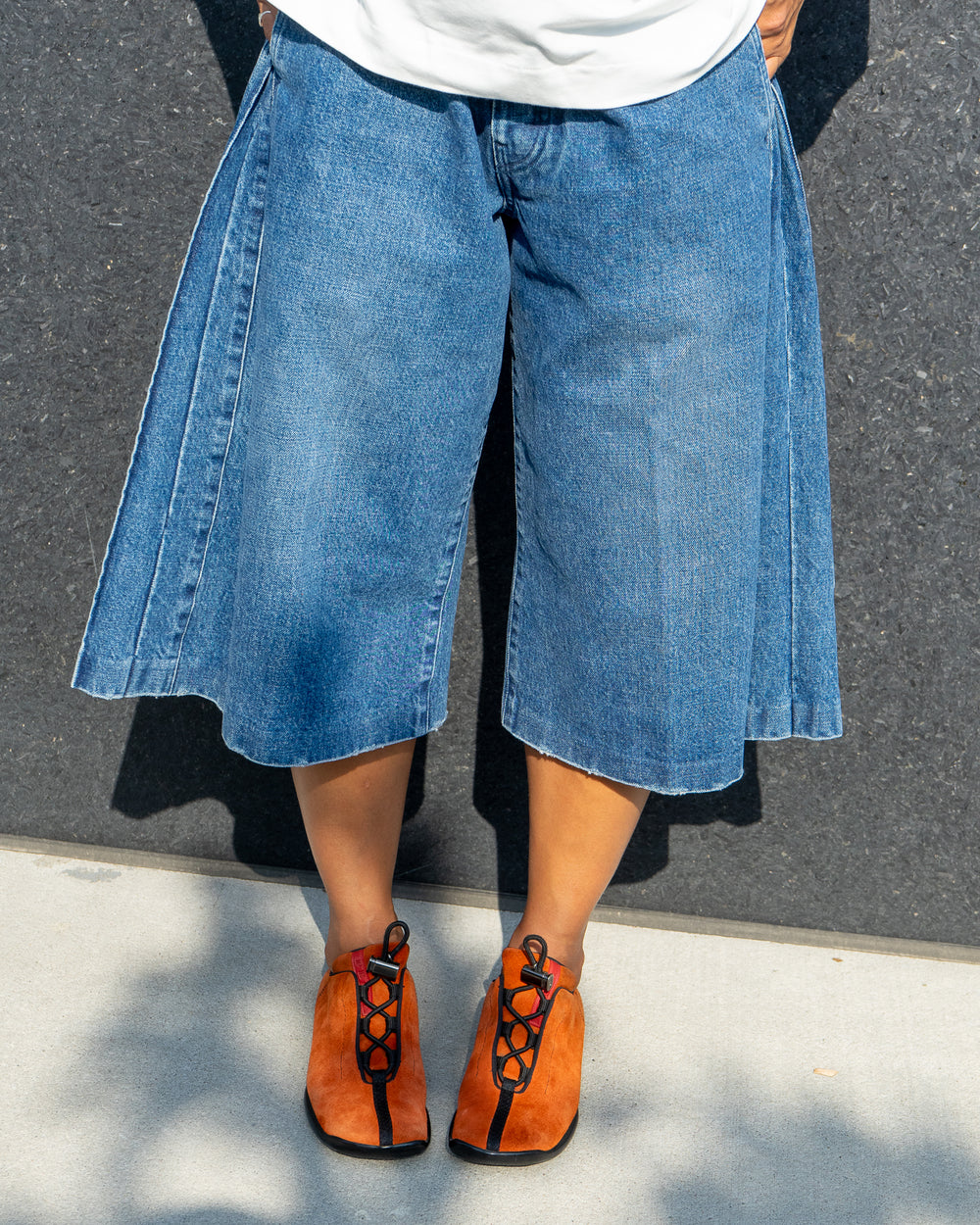 Person wearing blue denim culottes and orange shoes on a gray pavement.
