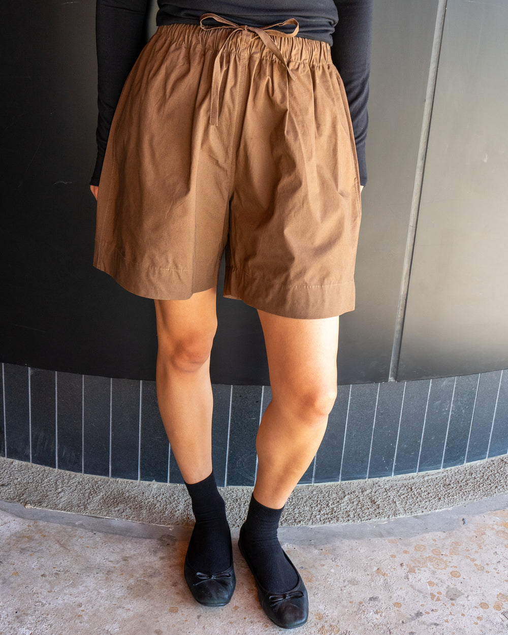 Person wearing brown shorts and black shoes against a tiled wall.